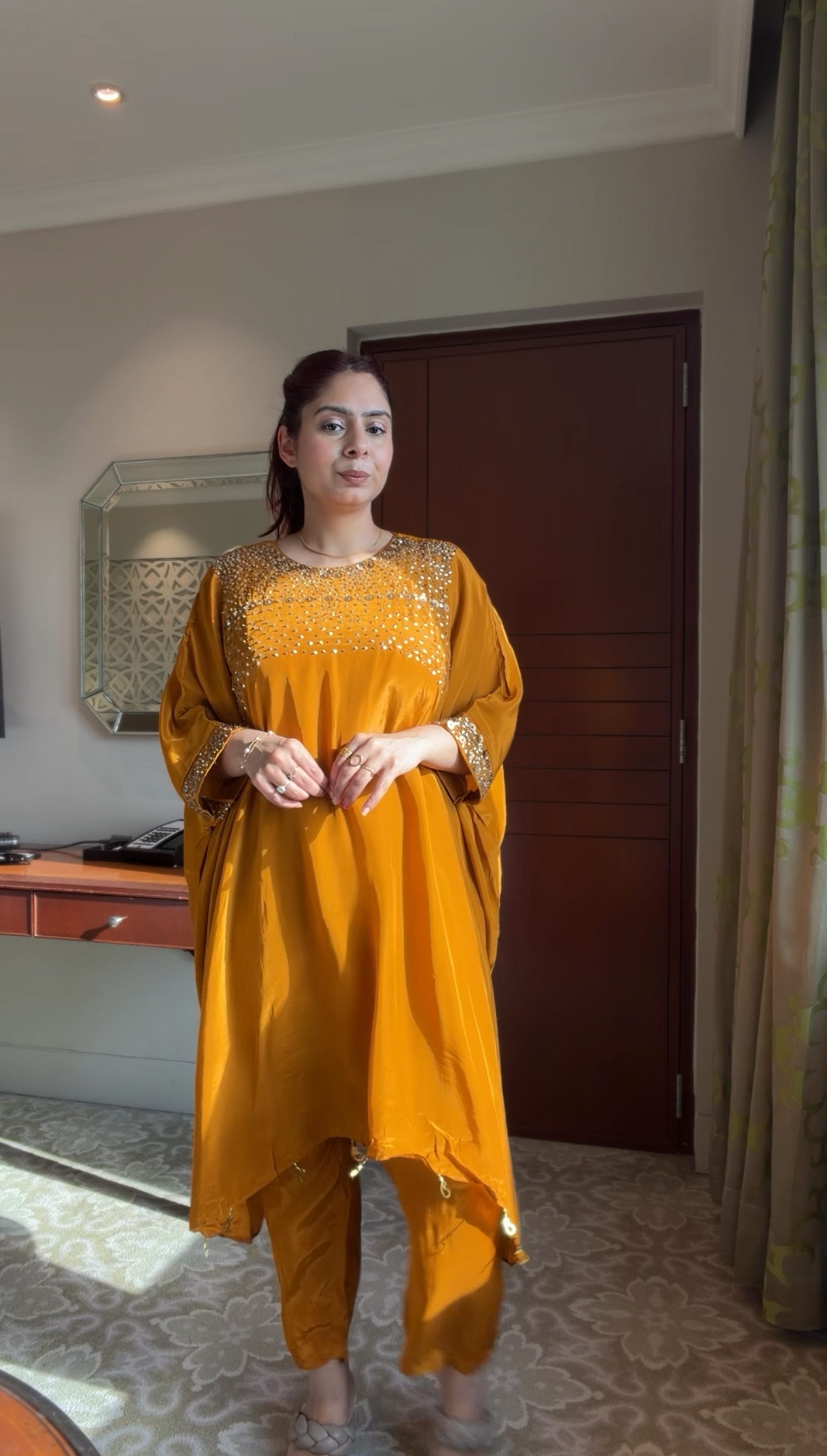 Woman wearing a mustard yellow traditional outfit standing in a room with a mirror and wooden door.